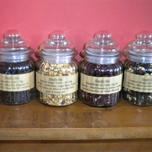 May include: Four glass jars with lids filled with loose leaf tea. The jars are labeled with the text "Totally Tea" and the names of the tea blends: Countess Grey, Lemon & Ginger Tango, Strawberries & Cream, and Lemongrass Orange Spice.