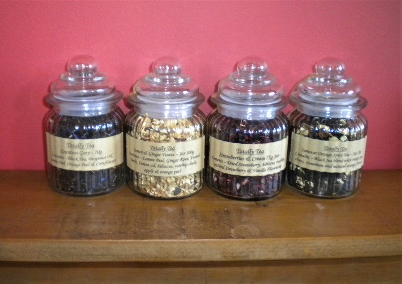 May include: Four glass jars with lids filled with loose leaf tea. The jars are labeled with the text "Totally Tea" and the names of the tea blends: Countess Grey, Lemon & Ginger Tango, Strawberries & Cream, and Lemongrass Orange Spice.
