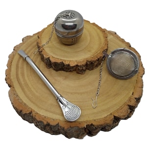 May include: A stainless steel spherical tea infuser with a chain, a mesh tea strainer with a chain and hook, and a metal bombilla straw with a perforated filter end. These three silver-toned tea accessories are arranged on a wooden surface.