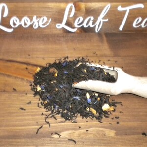 May include: Loose leaf tea with blue and orange pieces in a white scoop on a wooden surface.