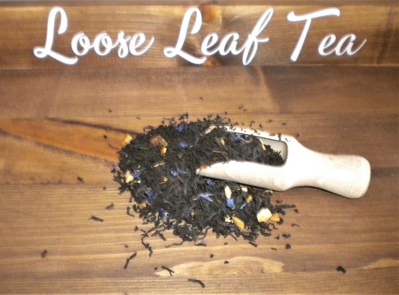 May include: Loose leaf tea with blue and orange pieces in a white scoop on a wooden surface.