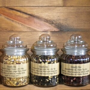 May include: Three glass jars filled with loose leaf tea blends. The jars have brown labels with the text "Totally Tea" and descriptions of the tea blends: "Lemon & Ginger Twine", "Summer Orange Spice Tea", and "Strawberries & Cream Tea".
