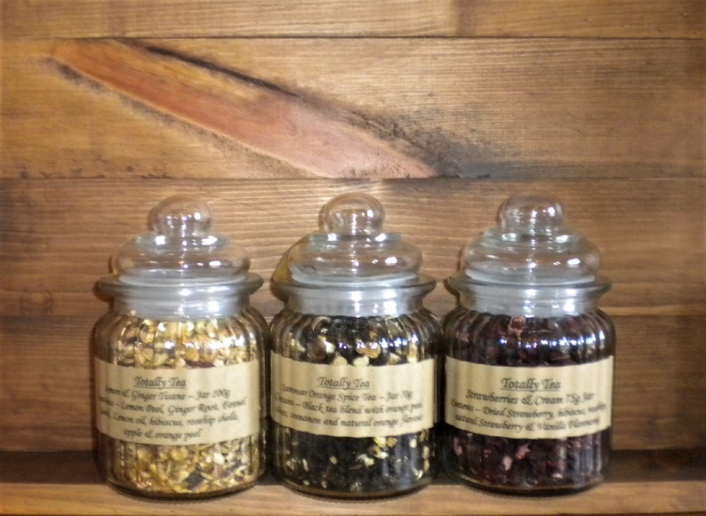 May include: Three glass jars filled with loose leaf tea blends. The jars have brown labels with the text "Totally Tea" and descriptions of the tea blends: "Lemon & Ginger Twine", "Summer Orange Spice Tea", and "Strawberries & Cream Tea".