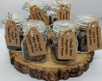 May include: Eight glass jars with brown paper tags tied with twine, each containing loose leaf tea. The jars are arranged on a wooden slice. The tags are labeled with the tea flavors: Jasmine Moonlight Green Tea with Jasmine, Masala India Spiced, Berry Burst Mixed Fruit, Totally Mint Peppermint Herbal Tisane, White Truffle White Tea & Coconut Blend, Morning Brew English Breakfast Tea.
