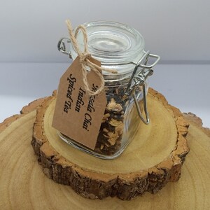 May include: A glass jar with a metal clasp lid filled with loose leaf Indian chai spiced tea. The jar is sitting on a wooden surface and has a brown paper tag tied around it with twine. The tag reads "Indian Chai Spiced Tea".