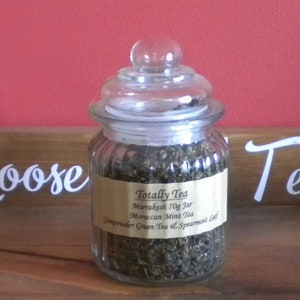 May include: A glass jar filled with loose green tea leaves. The jar has a glass lid and a label that reads "Totally Tea Marrakesh 70g Jar Moroccan Mint Tea Gunpowder Green Tea & Spearmint Leaf".