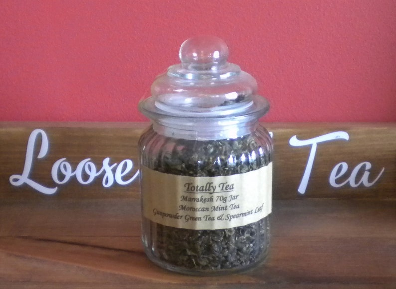 May include: A glass jar filled with loose green tea leaves. The jar has a glass lid and a label that reads "Totally Tea Marrakesh 70g Jar Moroccan Mint Tea Gunpowder Green Tea & Spearmint Leaf".