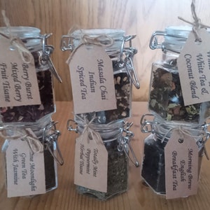 May include: Six glass jars with metal lids and brown paper tags. Each jar contains a different type of tea blend. The tags list the tea names: Berry Burst Mixed Berry Fruit Tisane, Masala Chai Indian Spiced Tea, Totally Mint Peppermint Herbal Tisane, White Truffle White Tea & Coconut Blend, Morning Brew English Breakfast Tea, and Fine Moonlight Green Tea With Jasmine.