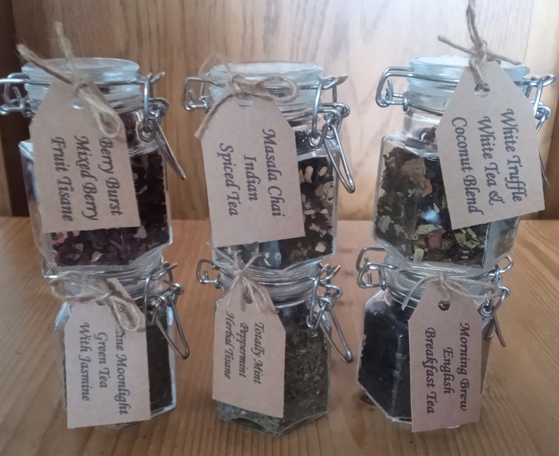 May include: Six glass jars with metal lids and brown paper tags. Each jar contains a different type of tea blend. The tags list the tea names: Berry Burst Mixed Berry Fruit Tisane, Masala Chai Indian Spiced Tea, Totally Mint Peppermint Herbal Tisane, White Truffle White Tea & Coconut Blend, Morning Brew English Breakfast Tea, and Fine Moonlight Green Tea With Jasmine.