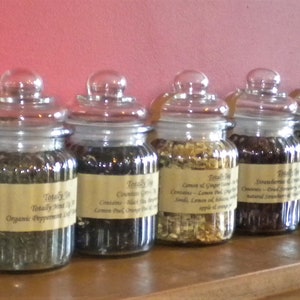 May include: Ten glass jars filled with loose leaf tea blends. The jars are labeled with the names of the tea blends, including "Christmas Cookie Fruit Tea", "Organic Peppermint", "Green Tea", "Lemon & Ginger", "French Earl Grey", "Jasmine Green", "Rooibos", "Chamomile", and "Peppermint".