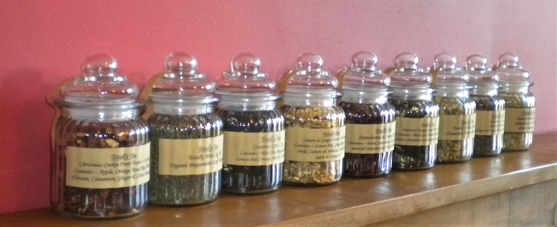 May include: Ten glass jars filled with loose leaf tea blends. The jars are labeled with the names of the tea blends, including "Christmas Cookie Fruit Tea", "Organic Peppermint", "Green Tea", "Lemon & Ginger", "French Earl Grey", "Jasmine Green", "Rooibos", "Chamomile", and "Peppermint".