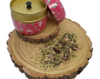 May include: A round metal tea tin with a pink floral pattern, a gold-coloured inner lid, and a small black knob. Beside the open tin, a pile of loose-leaf tea is visible, containing green tea needles, dried flower petals, and small red berries.