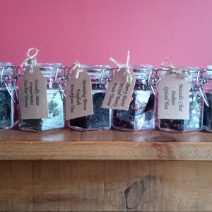 May include: Eight glass jars with brown paper tags, each containing a different type of loose leaf tea. The jars are arranged in a row on a wooden surface. The tags list the tea flavours: Jasmine Moonlight Green Tea, Totally Mint Peppermint Herbal Tisane, Morning Brew English Breakfast Tea, Berry Burst Mixed Berry Fruit Tisane, Indian Masala Chai Spiced Tea, White Truffle White Tea & Coconut Blend.