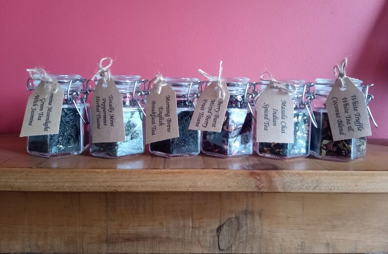 May include: Eight glass jars with brown paper tags, each containing a different type of loose leaf tea. The jars are arranged in a row on a wooden surface. The tags list the tea flavours: Jasmine Moonlight Green Tea, Totally Mint Peppermint Herbal Tisane, Morning Brew English Breakfast Tea, Berry Burst Mixed Berry Fruit Tisane, Indian Masala Chai Spiced Tea, White Truffle White Tea & Coconut Blend.