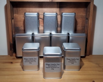 May include: Eight silver metal tea tins with black labels. The labels have the text "Mags Totally Tea" and the name of the tea variety. The tea varieties are Lapsang Souchong, Morning Brew, Himalayan Darjeeling.