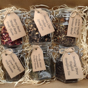 May include: A box containing six glass jars of loose leaf tea. Each jar has a brown paper tag with the name of the tea written on it. The teas are: White Truffle White Tea & Coconut Blend, Morning Brew English Breakfast Tea, Totally Mint Peppermint Herbal Tisane, Jasmine Moonlight Green Tea with Jasmine, Berry Burst Mixed Berry Fruit Tisane, and Masala Chai Indian Spiced Tea.