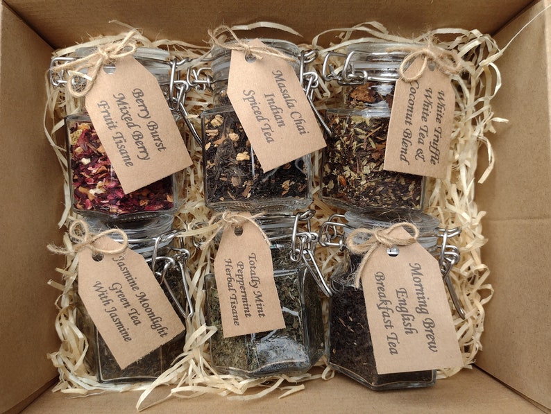 May include: A box containing six glass jars of loose leaf tea. Each jar has a brown paper tag with the name of the tea written on it. The teas are: White Truffle White Tea & Coconut Blend, Morning Brew English Breakfast Tea, Totally Mint Peppermint Herbal Tisane, Jasmine Moonlight Green Tea with Jasmine, Berry Burst Mixed Berry Fruit Tisane, and Masala Chai Indian Spiced Tea.