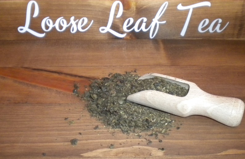 May include: A wooden scoop filled with loose leaf tea on a wooden surface. The tea is a dark green color and has a rough texture. The text "Loose Leaf Tea" is visible on the surface above the tea.