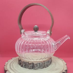 Puede incluir: Una tetera de vidrio transparente con diseño acanalado, asa de madera y pomo de madera en la tapa. La tetera está sobre una rodaja de madera sobre un fondo rojo. Diseñada para preparar té.