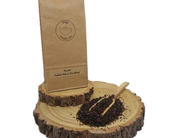 May include: A brown paper bag of Assam Indian Black Tea Blend with a circular logo featuring a teapot and the text Mags Totally Tea. A small wooden scoop rests on a pile of loose black tea leaves placed on a round, natural wood slice base.