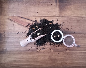 May include: A wooden scoop filled with black tea leaves and a white tea infuser with black tea leaves inside. The tea leaves are scattered on a brown wooden surface.