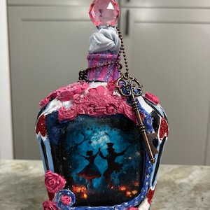 May include: Decorative bottle with a pink crystal stopper and a key charm. The bottle features a painted scene of dancing figures in a forest, with pink and blue accents, and decorative roses. The bottle is a unique decorative item.