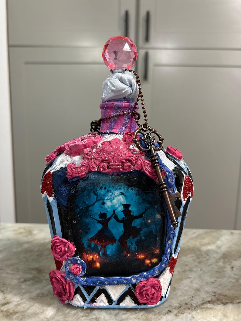 May include: Decorative bottle with a pink crystal stopper and a key charm. The bottle features a painted scene of dancing figures in a forest, with pink and blue accents, and decorative roses. The bottle is a unique decorative item.
