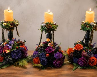 May include: Three black candle holders, each topped with a lit yellow pillar candle and a decorative ring of green moss and small metallic spheres. The base of each holder is adorned with a dense arrangement of purple, orange, and black artificial roses.