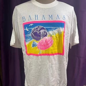May include: White t-shirt with a colorful graphic featuring the word "BAHAMAS" above a beach scene. The graphic includes a pink seashell, a purple seashell, and a yellow and blue beach background.