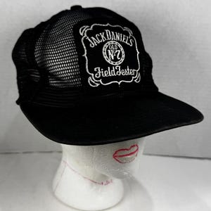 May include: A black trucker hat featuring a mesh back and a solid front panel. The front displays a white embroidered patch with the text Jack Daniel's Old No. 7 Field Tester in a decorative frame. The hat has a flat brim and a structured crown.
