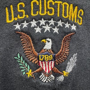 Può includere: Un tessuto grigio scuro con un logo ricamato U.S. CUSTOMS in testo giallo sopra una fila di stelle bianche. Sotto le stelle c'è un'aquila calva con ali marroni, che tiene un ramo verde e delle frecce, con uno scudo che riporta l'anno 1789.