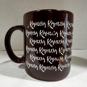 May include: A dark brown coffee mug with the word "KAHLUA" repeated in white script. The mug has a large handle and is likely made of ceramic. The mug is designed for hot beverages.