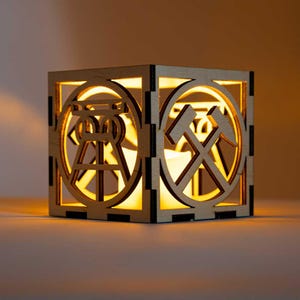 Könnte beinhalten: Ein quadratischer Kerzenhalter aus Holz mit ausgeschnittenen Designs. Der Kerzenhalter hat ein kreisförmiges Design mit Hammer und Spitzhacke und ein weiteres mit einem Turm. Das Innere leuchtet mit warmem Licht und schafft eine gemütliche Atmosphäre.