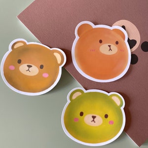 May include: Three round stickers featuring cartoon bear faces. The bears have brown, orange, and green fur. Each bear has a small pink blush on its cheeks.