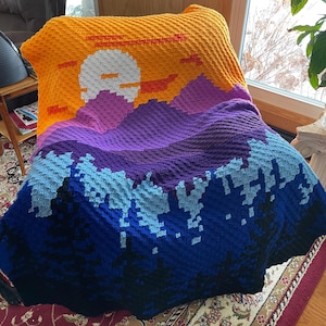 Peut inclure: Une couverture au crochet avec une scène de montagne pixélisée. La couverture représente un coucher de soleil avec un soleil blanc, des montagnes orange et rouges, des collines violettes et des arbres bleus.
