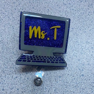 Op de afbeelding: Een blauwe glittercomputer met een gele glitter "Ms. T" op het scherm. De computer is bevestigd aan een intrekbare badgehouder met twee zilveren clips.