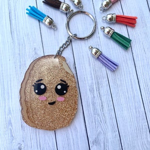 Può includere: Un portachiavi con glitter dorati e un designo a cartone animato di una patata fritta. Il portachiavi ha un anello argentato e viene fornito con una varietà di nappe colorate.