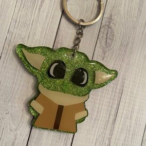 May include: Green glitter keychain featuring a cartoon character with large eyes and brown clothing.