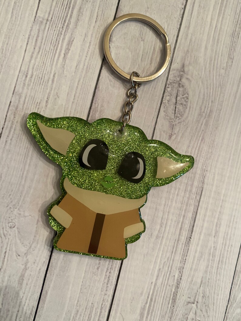 May include: Green glitter keychain featuring a cartoon character with large eyes and brown clothing.