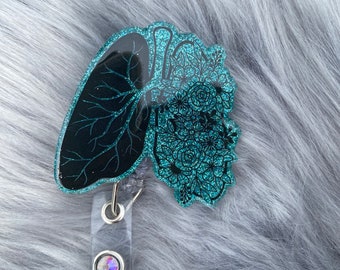 May include: Teal blue glitter resin badge reel featuring a black and teal anatomical heart and lung design with floral accents. The reel has a clear plastic strap and a sparkly iridescent gem.