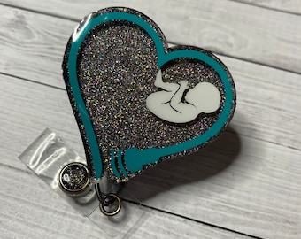 May include: A teal blue heart-shaped badge holder with silver glitter and a white silhouette of a baby inside. The badge holder has a clear plastic clip.