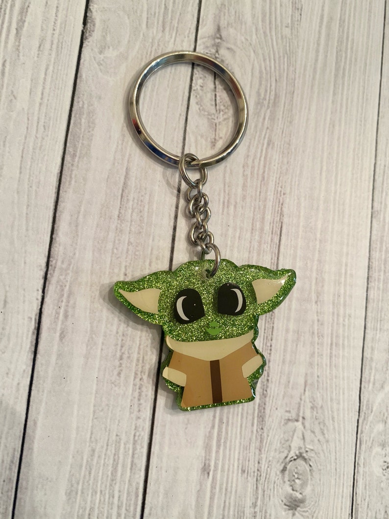 May include: A green glitter keychain featuring a cartoon character with large eyes. The character is wearing a brown shirt and has large ears.
