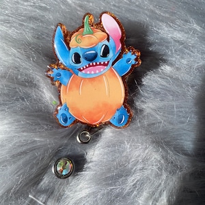 May include: A blue and orange glitter badge holder featuring a cartoon character dressed as a pumpkin. The character has large eyes and a wide smile. The badge holder has a clear plastic backing and a silver metal clip.