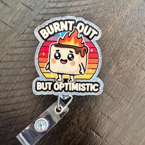 May include: A badge reel with a cartoon marshmallow character on a colorful sunset background. The text reads "BURNT OUT BUT OPTIMISTIC". The reel has a clear plastic strap with a decorative iridescent button.