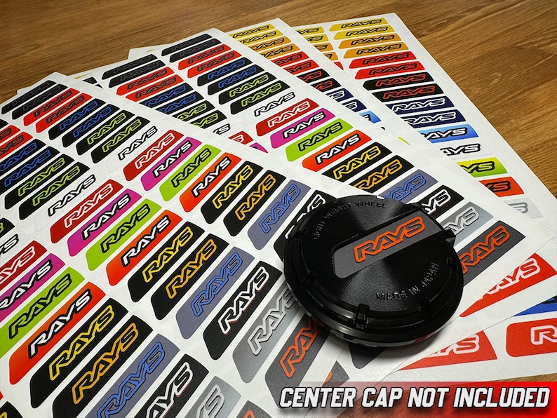 May include: A set of colorful stickers with the word "RAYS" printed on them. The stickers are arranged in rows on a white background. A black center cap with the word "RAYS" printed on it is in the foreground. The text "CENTER CAP NOT INCLUDED" is printed at the bottom of the image.