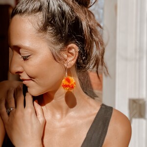 Può includere: Una donna indossa una canotta nera con un paio di orecchini a uncinetto arancioni e gialli. Gli orecchini sono rotondi e hanno una superficie testurizzata.