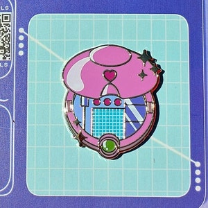 May include: A pink and white mushroom-shaped enamel pin with a heart and star design, set on a blue card with a grid pattern. The card has a QR code and social media icons. The pin is approximately 1.5 inches in diameter.