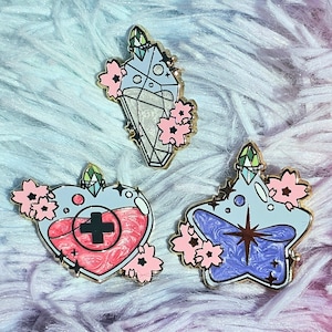 May include: Three enamel pins featuring different potion bottles with floral accents. The first pin is a blue crystal-shaped bottle with a gold top and pink flowers. The second pin is a red heart-shaped bottle with a gold top and pink flowers. The third pin is a blue star-shaped bottle with a gold top and pink flowers.