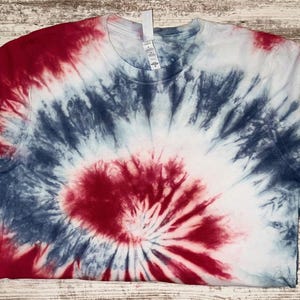 Pode incluir: Uma camiseta de manga curta com um padr&atilde;o tie-dye em vermelho, branco e azul. O design irradia do centro, criando um efeito espiral. A camisa &eacute; feita de um material macio.