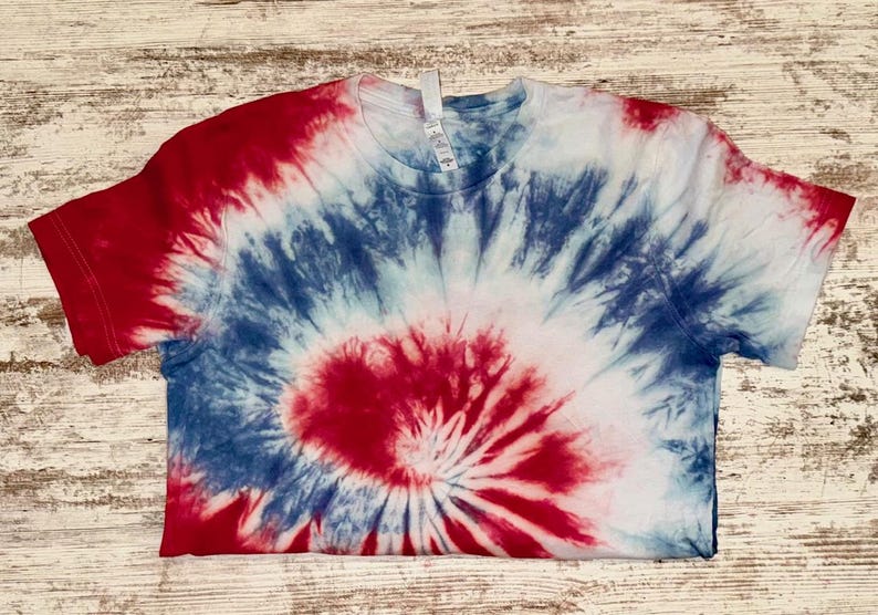 Pode incluir: Uma camiseta tie-dye vermelha, branca e azul. A camiseta de manga curta apresenta um design em espiral, com vermelho no centro, passando para azul e branco. A camiseta est&aacute; colocada em uma superf&iacute;cie de madeira clara.
